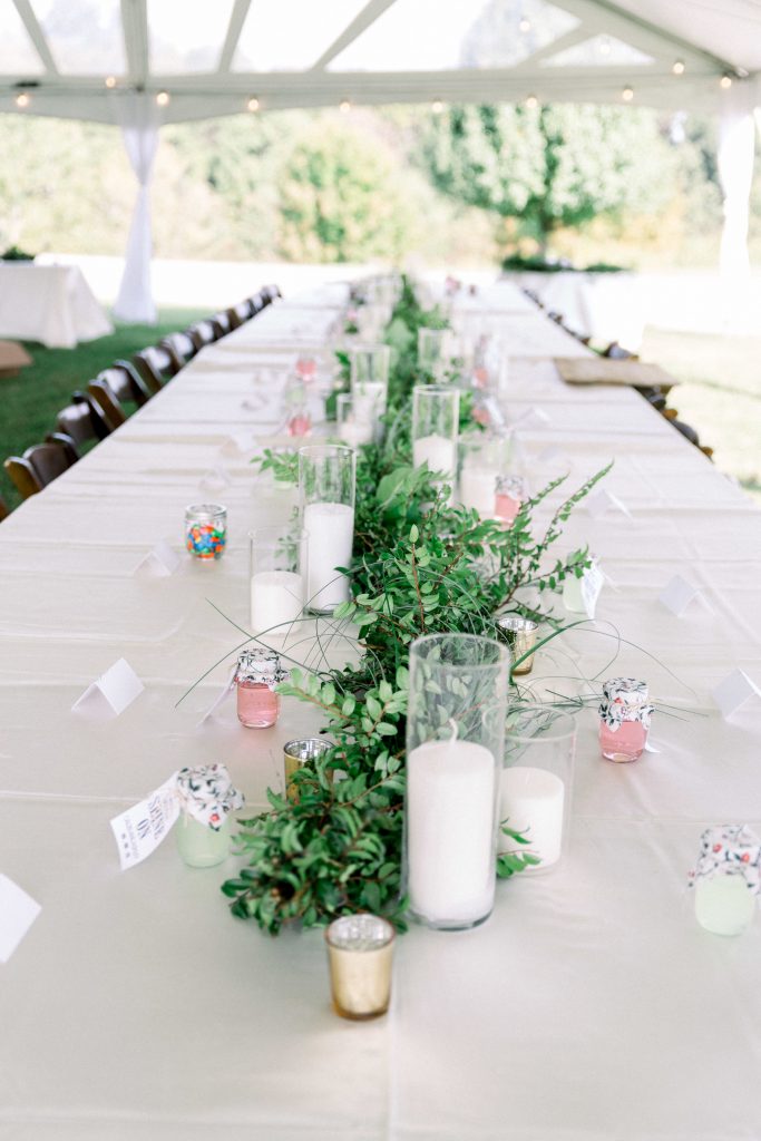 Table decor, on long white covered tables. Greenery runs down the middle with different height white candles alternating on each side of the greenery.
