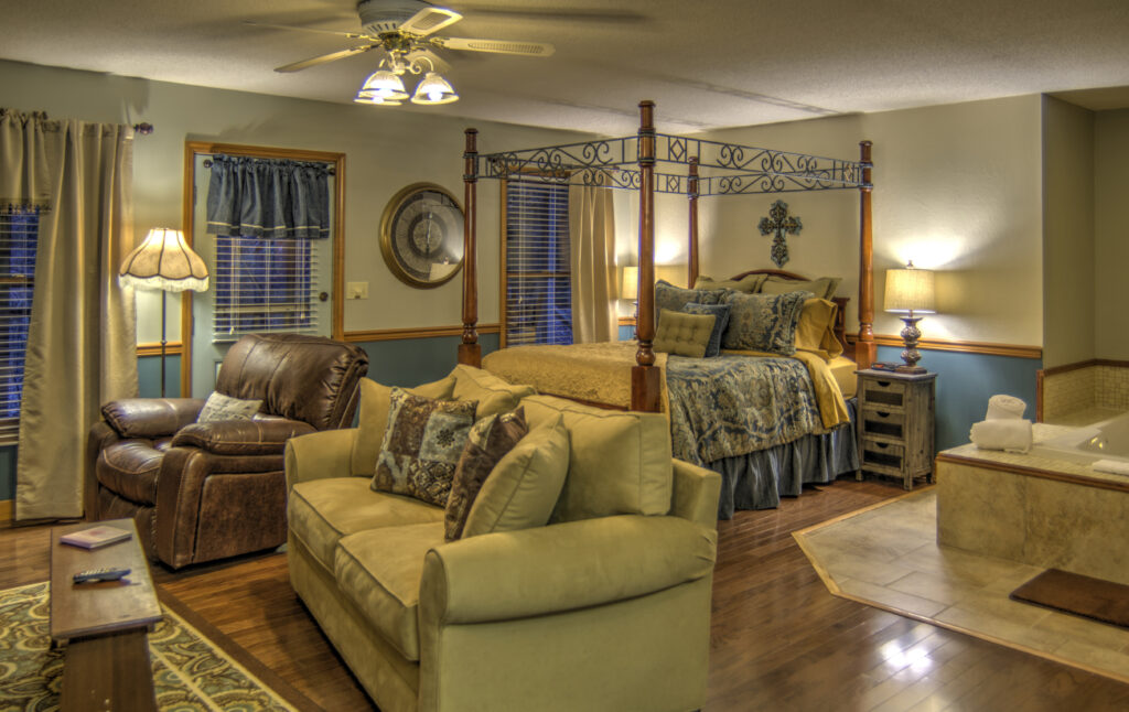 A wide view of a suite with a 4-poster bed, sofa, recliner, and jacuzzi tub.