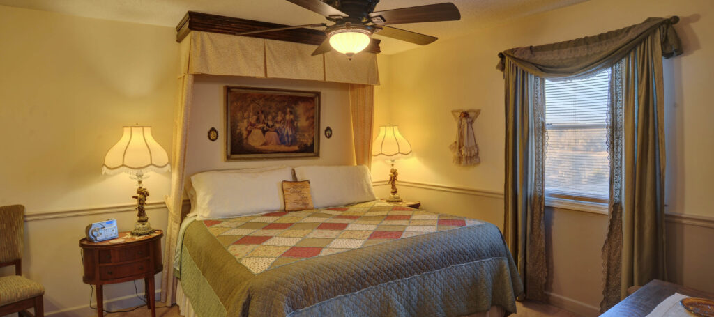 A wide view of a hotel room with a bed, nightstand, lamp, ceiling fan, and window.