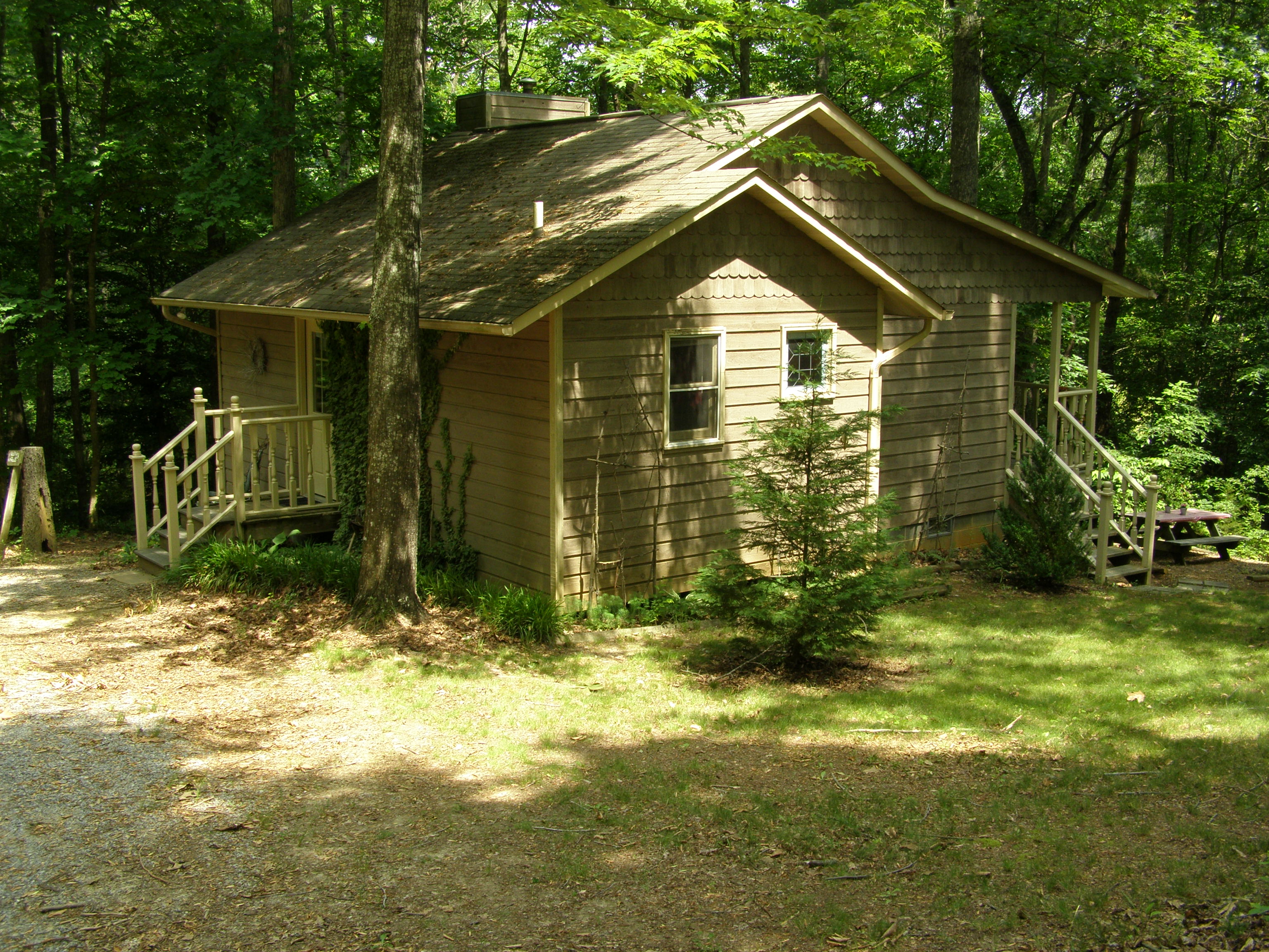 A small, brown, wooden cottage with a porch and stairs, surrounded by trees and a grassy area.